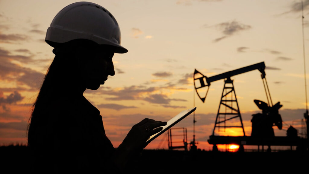 Silueta de una operaria petrolera frente a bombas de extracción de petróleo al atardecer.